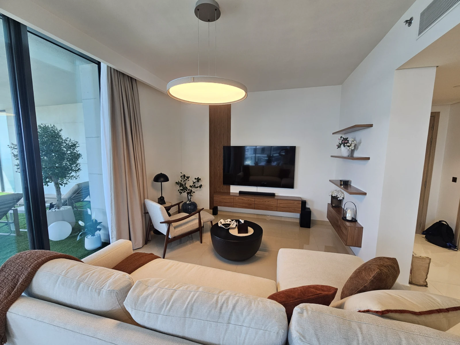 A Donna Branka living room at Bay Residence: a slatted wood panel behind the TV, floating walnut shelves, a ring pendant and a terrace with an olive tree.